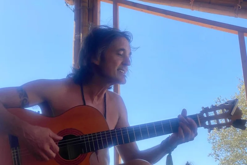 Manu tocando la guitarra bajo un techo de madera, con el cielo azul detrás